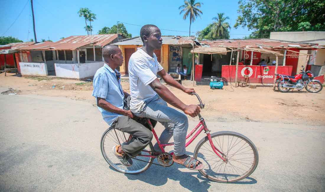 Zwei Männer in Hemd, Hose und Sandalen überqueren in Mosambik auf einem roten Fahrrad einen leeren Platz. Im Hintergrund überdachte Marktstände und ein geparktes Motorrad.