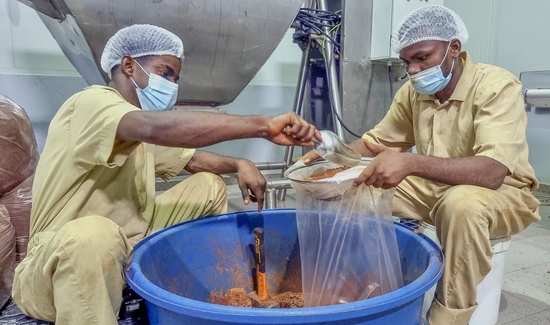 Zwei sitzende junge angolanische Fabrikarbeiter in heller Arbeitskleidung, mit Mundschutz und Haarhaube, füllen mit einer Metallschaufel Kakao aus einem großen blauen Bottich in eine Metallschüssel. 
