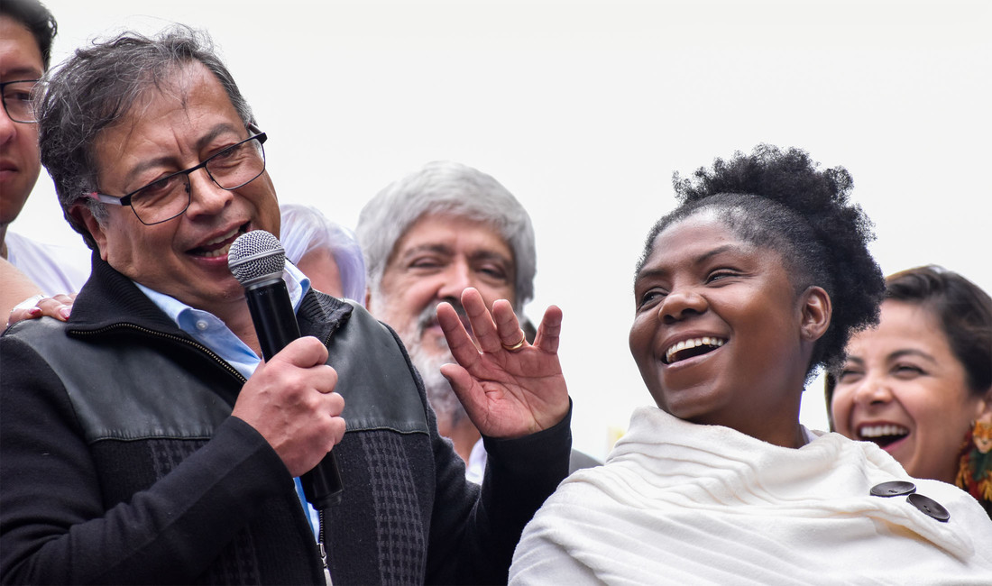 Nahaufnahme des grauhaarigen kolumbianischen Präsidenten Gustavo Petro mit Mikrofon in der Hand, neben ihm die lachende schwarzhaarige afrokolumbianische Vizepräsidentin Francia Márquez, im Hintergrund fröhliche Gesichter. 