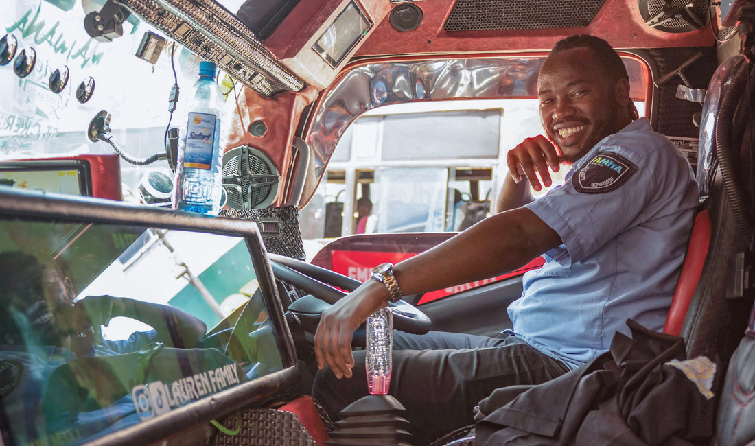 Ein strahlend lachender Afrikaner, Charles Gachunji, im kurzärmeligen hellblauen Hemd einer Busfahrer-Uniform auf dem Fahrersitz hinter dem Steuer eines Busses. Der Bus hat eine rote Innenverkleidung.