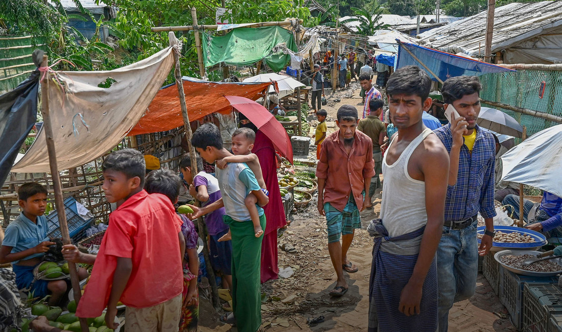 Rohingya-Männer und -Jungens drängen sich im Flüchtlingslager zwischen mit löchrigen Tüchern und Schirmen vor der Sonne geschützten provisorischen Obst- und Gemüseständen. Ganz links im Bild ein kleiner Junge in blauen Shorts, der gelbgrüne Früchte verkauft.