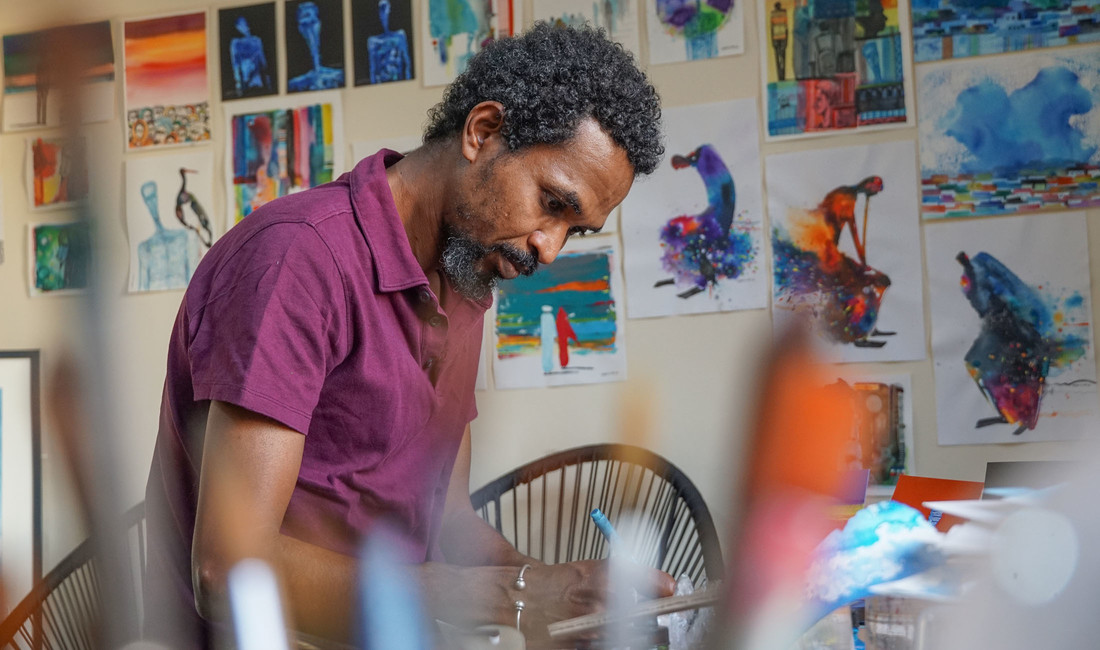 Halbporträt eines sudanesischen Künstlers mittleren Alters im Profil. Der bärtige Mann im kurzärmeligen rotvioletten Hemd beugt sich über ein Zeichenbrett. Die Atelierwand hinter ihm ist gepflastert mit farbenfrohen gemalten Bildern.