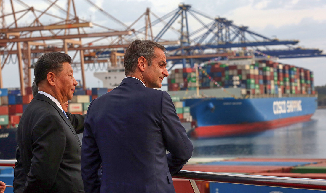 Chinas Präsident Xi Jinping und Griechenlands Premierminister Kyriakos Mitsotaki, beide in dunklen Anzügen und von hinten im Profil zu sehen, blicken auf ein Hafenbecken mit beladenen Containerschiffen.