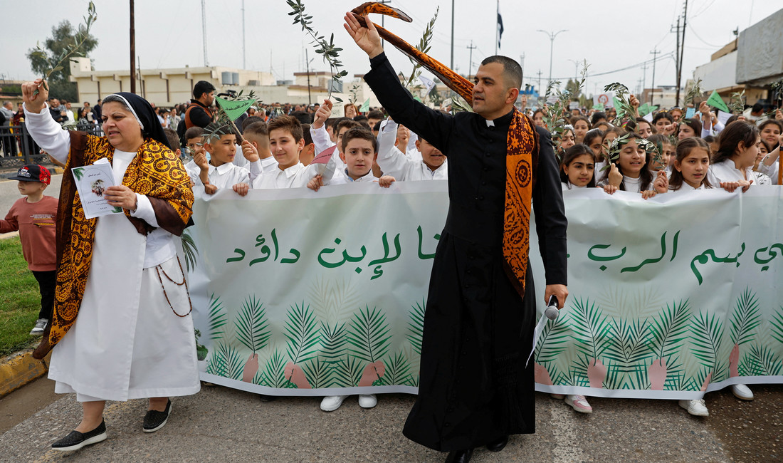 Ein schwarze gewandeter Priester und eine Nonne führen eine Prozession an, vor einem arabisch beschrifteten Banner.