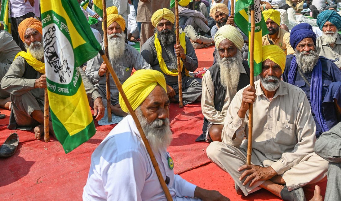 Protestdemonstration indischer Bauern. Traditionell gekleidet, mit Bärten und farbenfrohen Turbanen, sitzen sie am Boden, Fahnenstangen in den Händen haltend.