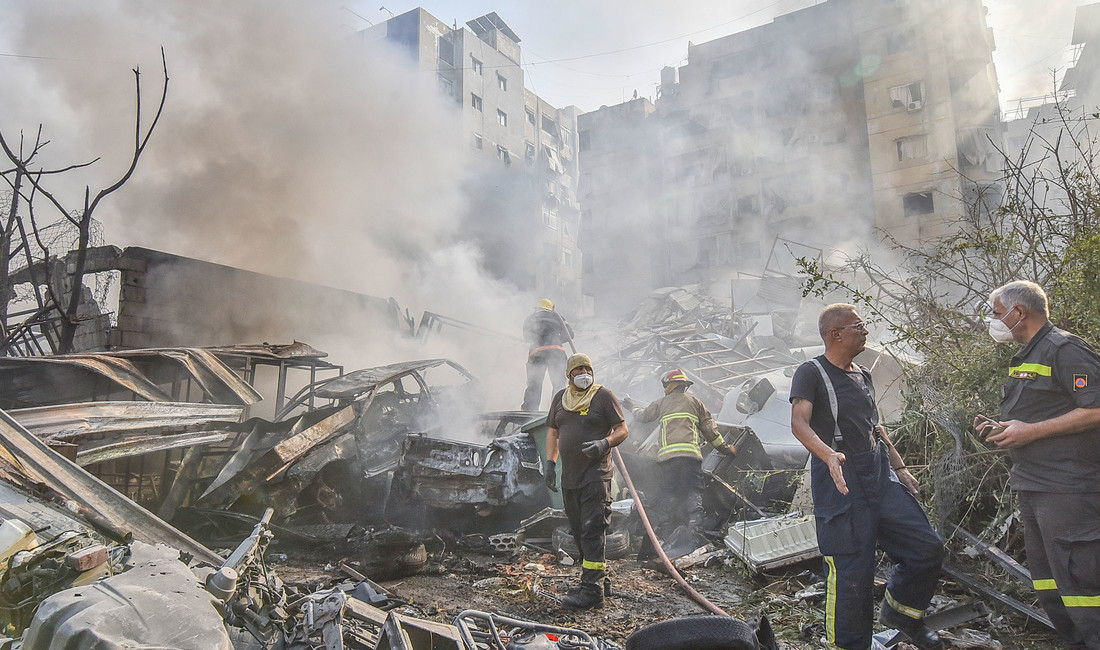 Fünf Feuerwehrmänner, zwei davon mit Mundschutz, einer mit einem langen Schlauch, in dichtem Qualm vor zerbombten Häusern eines Vororts von Beirut. Links im Bild Autowracks