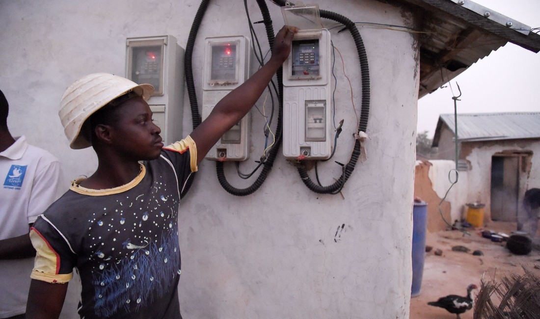 Ein junger Afrikaner deutet auf zwei Stromzähler einer Solaranlage, die an einer Hauswand befestigt sind. 