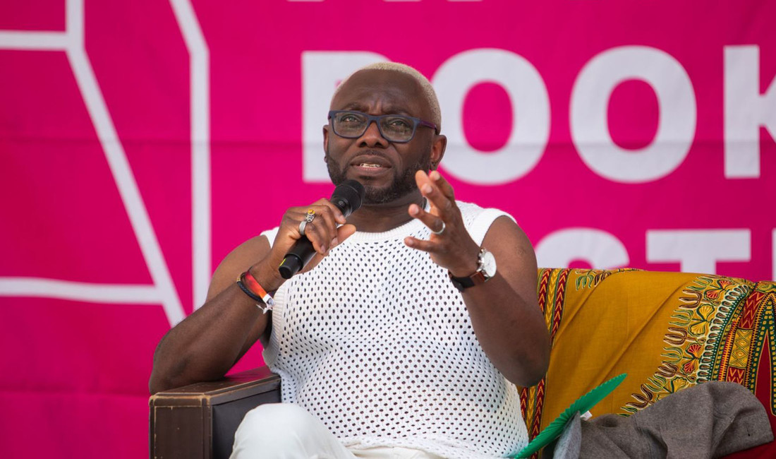 Ein nigerianischer Schriftsteller mit ärmellosem weißem Shirt, Brille und Mikrofon in der Hand im Halbporträt vor einem magentafarbenen Hintergrund. 