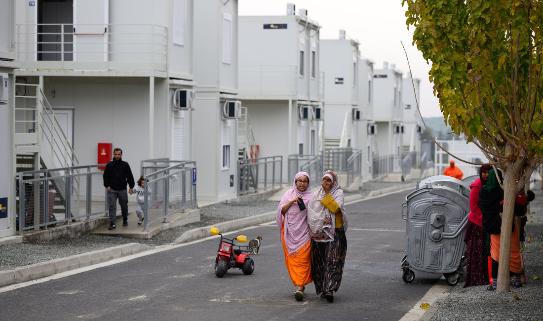 Im Hintergrund stehen mehrere Flüchtlingscontainer an einer Straße, auf der zwei afrikanische Frauen laufen. 