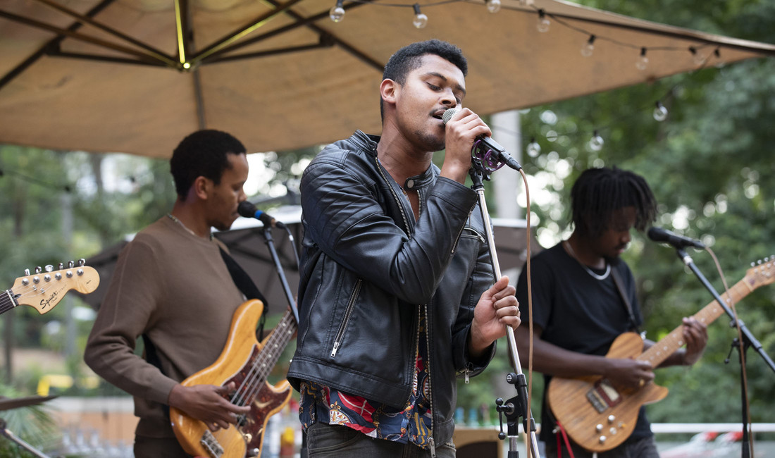 Drei Mitglieder einer kenianischen Punkrockband bei einem Auftritt auf der Bühne im Halbporträt; im Mittelpunkt und Vordergrund ein junger Sänger in Lederjacke am Mikrofon. 