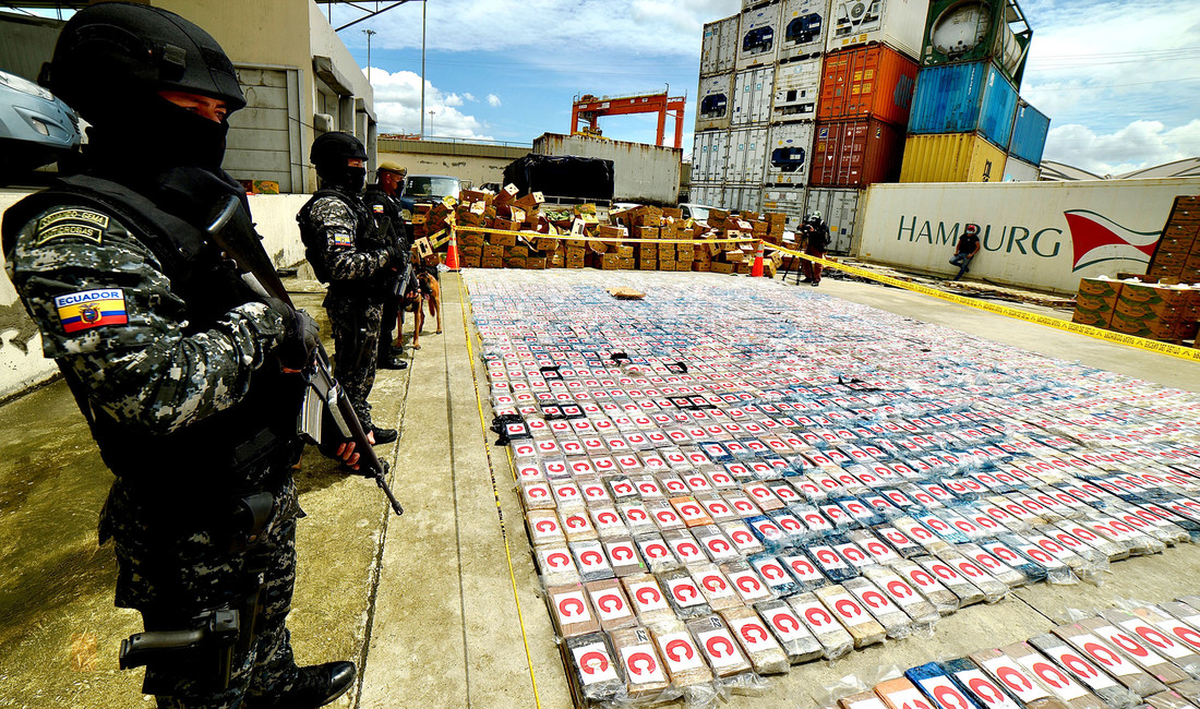 Polizisten mit gefleckten Uniformen, schwarzen Helmen, Westen und Stiefeln sowie Gewehren in der Hand bewachen im Hafen von Guayaquil auf dem Boden ausgebreitete Kokain­päckchen.