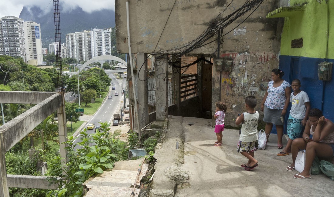 Auf der rechten Bildseite zwei Frauen, ein Junge im Teenageralter, ein kleiner Junge und ein kleines Mädchen vor einem hässlichen nackten Betonbau in einem brasilianischen Armenviertel. Sie blicken von einer Anhöhe herab auf eine Straße, auf der Autos fahren. Auf der linken Seite des Bildes sieht man in nicht allzugroßer Ferne weiße Hochhäuser in einer Grünanlage mit Bäumen.