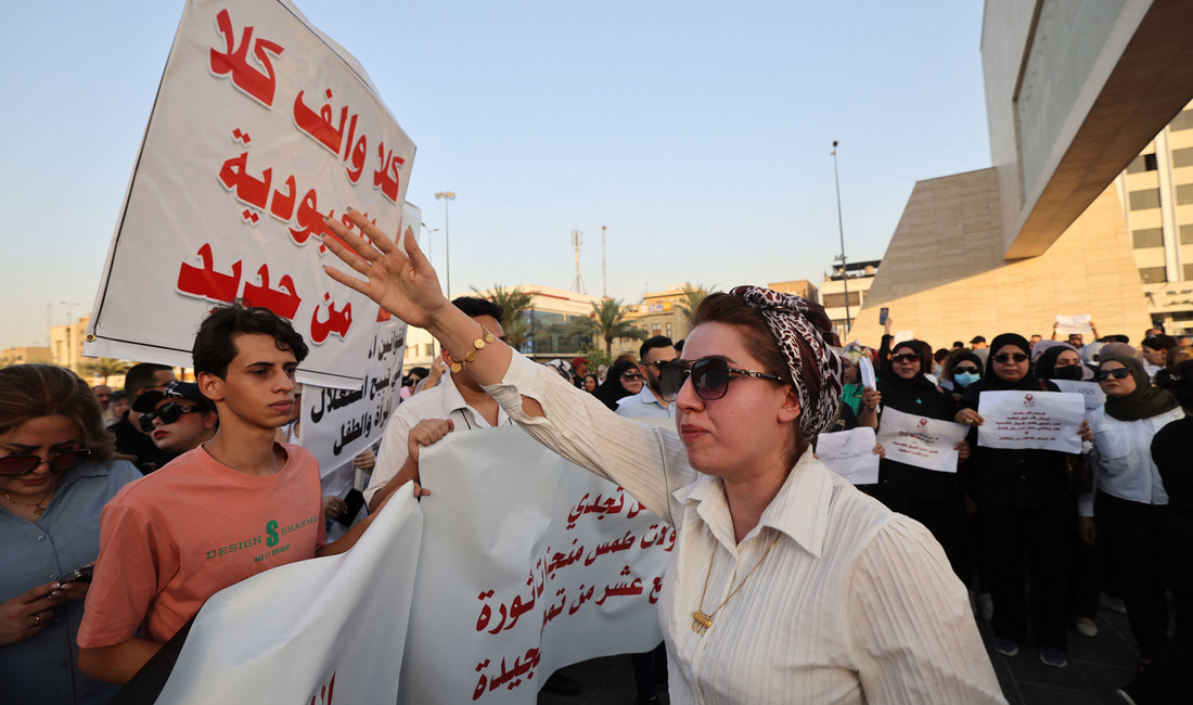 Eine Irakerin in weißer Bluse und mit Sonnenbrille, den Arm erhoben, vor der Kulisse einer Protestdemonstration. Manche der Frauen im Hintergrund sind schwarz gekleidet und tragen Schilder vor sich her. Links im Bild auch Männer.