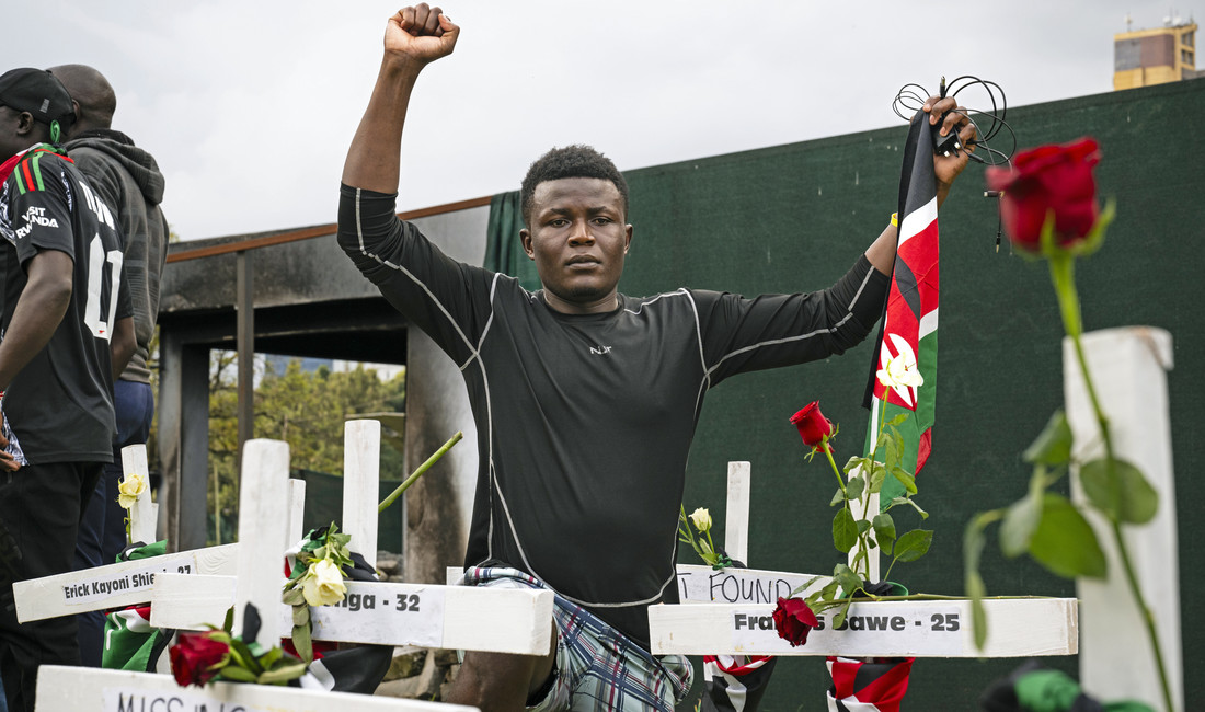 Ein junger Mann kniet zwischen Kreuzen mit Rosen, eine Faust geballt, in der anderen Hand eine Fahne Kenias.