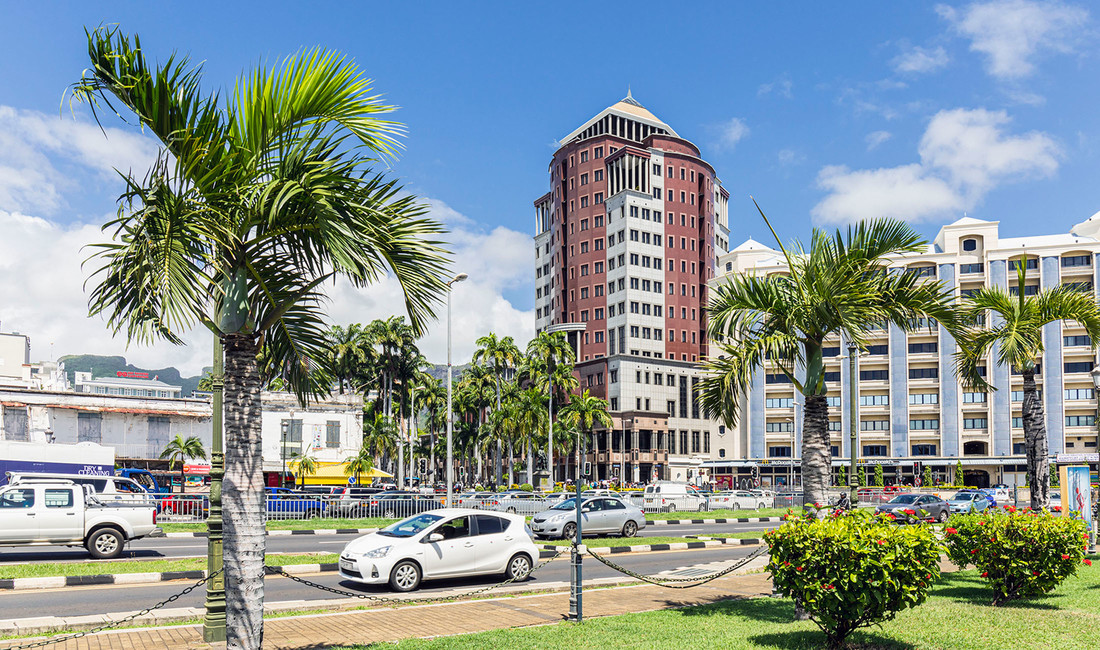 Ein Tower zwischen modernen Häusern in Port Louis, Mauritius. Im Vordergund eine mehrspurige Straße befahren von Autos, davor ein Rasenstreifen mit Palmen.