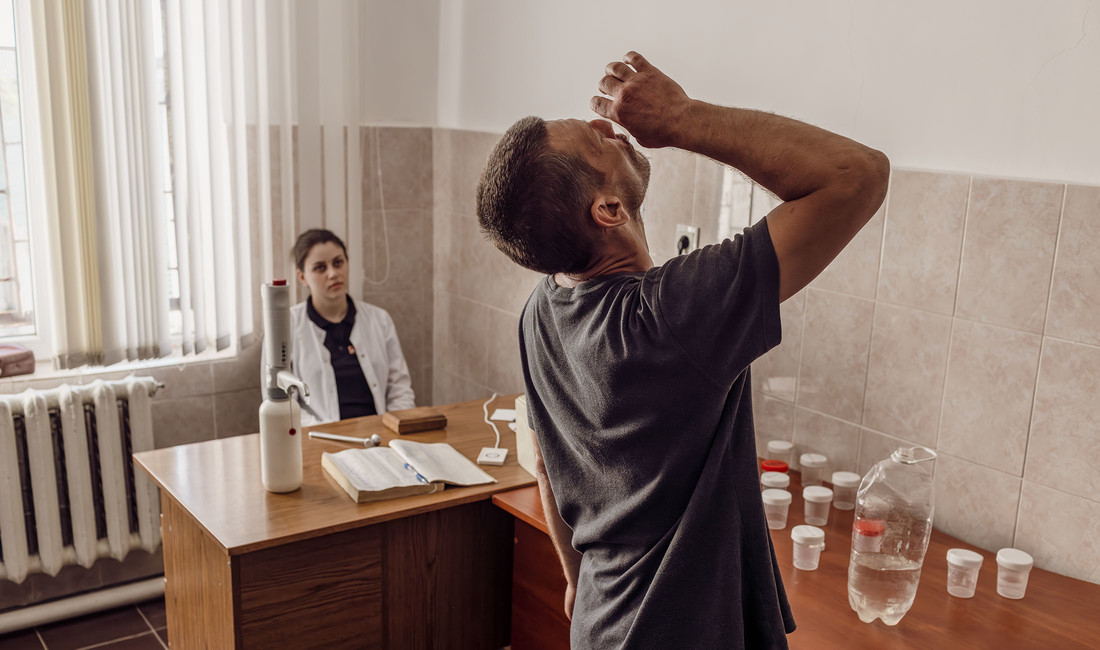 Ein Mann in grauem T-Shirt, von hinten gesehen, schluckt im Rahmen eines Drogenentzugs ein in Wasser aufgelöstes Medikament. Er streht vor einem Tisch mit Tabletten-Döschen und einem Wasserbehälter. Im Hintergrund eine Gefängnisärztin in weißem Kittel.