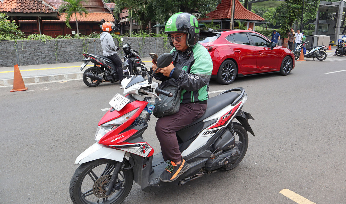 Indonesier mit grün-schwarzem Helm, grün-schwarzer Jacke und Turnschuhen auf einer weiß-roten Honda, hinter ihm ein rotes parkendes Auto, Menschen am Straßenrand, eine Mauer und ein parkähnliches Gelände