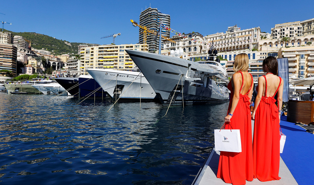 Zwei junge Frauen in rückenfreien langen roten Keidern vor Luxusschiffen im Hafenbecken des Yachthafens von Monaco.