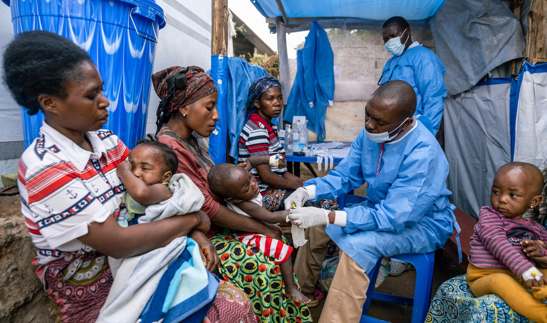 Frauen mit kleinen Kindern auf dem Schoß warten im Ostkongo auf eine Impfung gegen Mpox; ein afrikanischer Arzt mit Mundschutz, blauem Plastikkittel und weißen Handschuhen verabreicht einem der Kinder den Impfstoff.