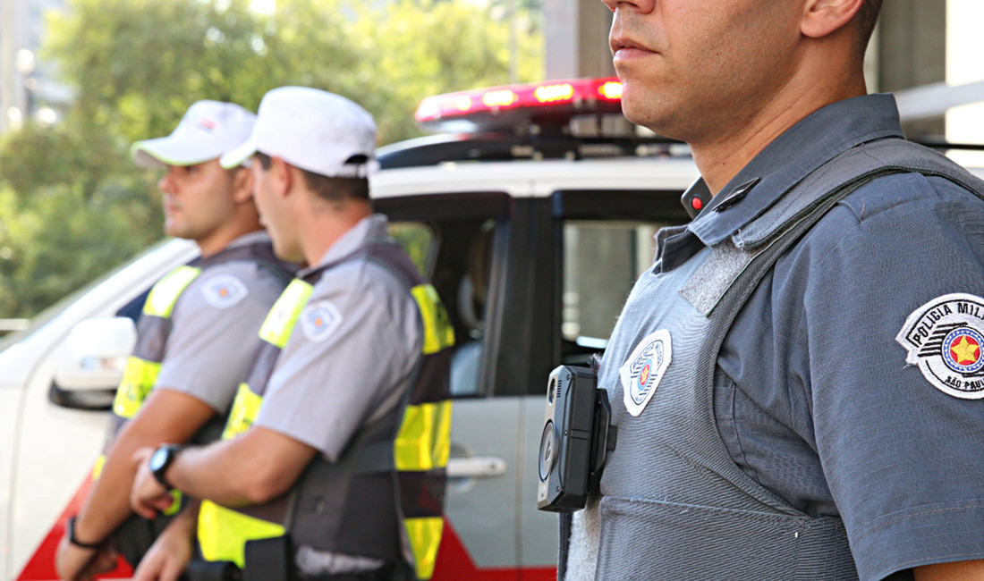 Halbporträt von drei Männern in Uniformen der brasilianischen Militärpolizei im Profil, die Gesicher sind nicht zu erkennen. Der Polizist, der rechts groß im Bild ist, trägt an seiner Weste in Brusthöhe eine Boycam.