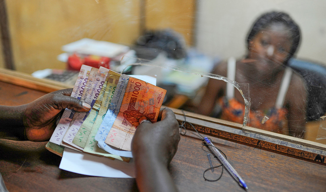 Hände mit verschiedenfarbigen Geldscheinen in CFA-Francs an einem Auszahlungsschalter mit Glasscheibe; dahinter verschwommen Gesicht und Schultern einer Afrikanerin 