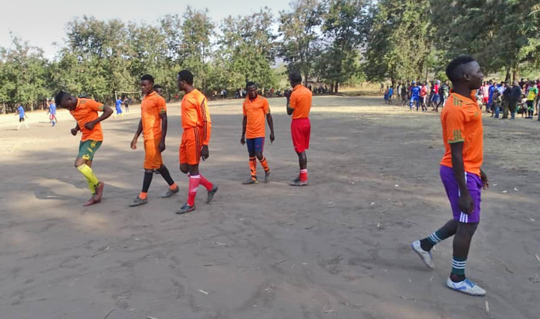 Sechs Schüler in orangenen Fußballtrikots auf einem Platz in Tansania. Im Hintergrund Bäume und Zuschauer.