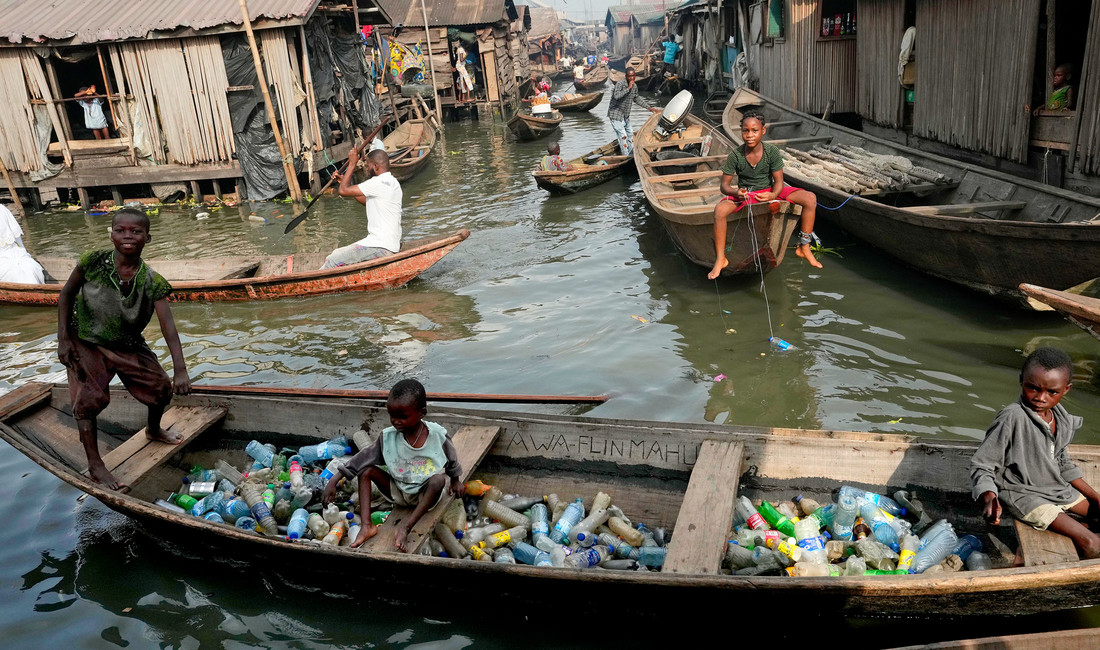 Im Vordergrund drei Jungen, zwei der Kinder sichtlich unterernährt, in einem Holzboot voller leerer Plastikflaschen. Dahinter weitere Boote mit Insassen. Sie fahren zwischen dicht gedrängten Slumhütten, die in bräunlich-grauem Wasser stehen.