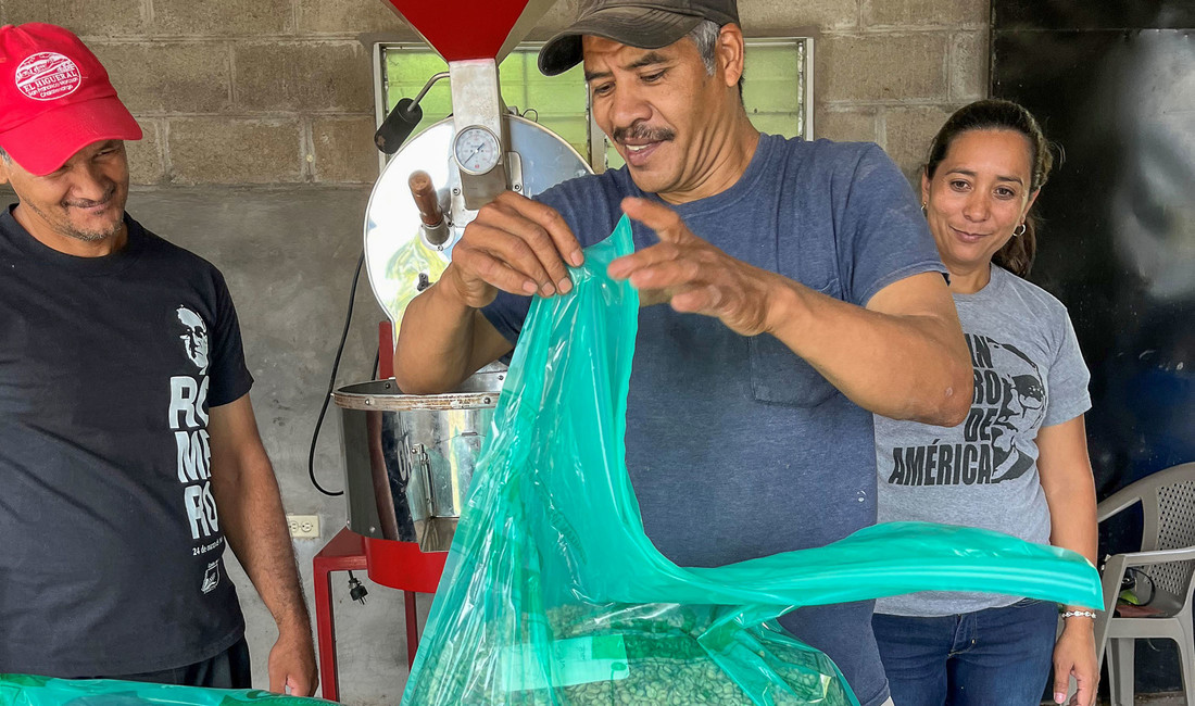 Arnoldo Arévalo, ein 50-jähriger Bauer in El Salvador mit Schnurrbart, schwarzer Schirmkappe und grauem T-Shirt, schaut zufrieden lächelnd auf einen durchsichtigen grünen Plastiksack mit Kaffeebohnen. Hinter ihm ein Mann mit roter Schirmkappe und eine dunkelhaarige Frau, die ebenfalls zufrieden auf die Kaffeeernte blicken.