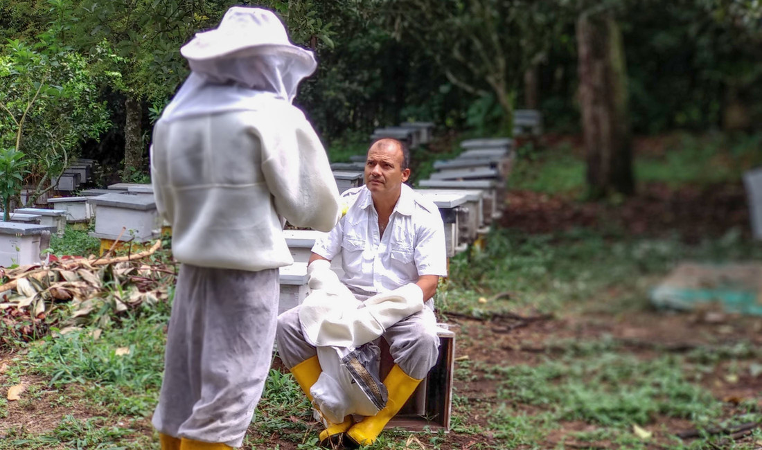 Zwei Männer in weißer Schutzkleidung für Imker und mit gelben Gummistiefeln unterhalten sich auf einem Gelände mit Bienenkästen in Kolumbien. 